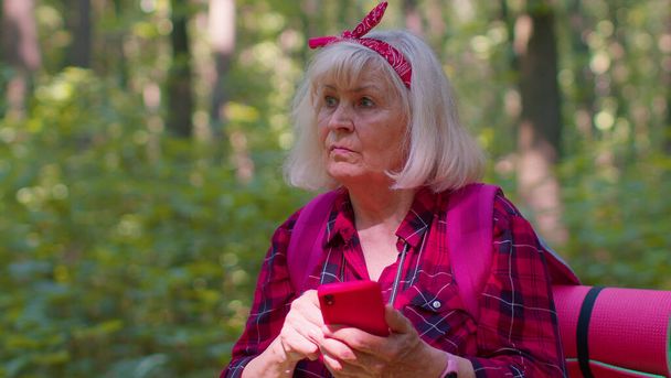 Çekici yaşlı büyükanne kadın sırt çantalı. Ormanda cep telefonuyla yolculuk planı yapıyor. - Fotoğraf, Görsel