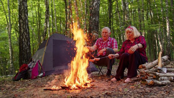 Senior joyeuse vieille grand-mère grand-père cuisine des saucisses frites sur le feu de camp dans le bois au camping - Photo, image