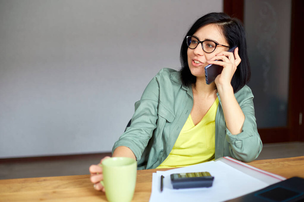 Stilvolle brünette Frau in Gläsern, die am Holztisch mit Notizblock sitzt und telefoniert, Tee trinkt am modernen Arbeitsplatz, freiberuflich zu Hause arbeitet. Kaffeepause im Büro, Ruhezeit - Foto, Bild