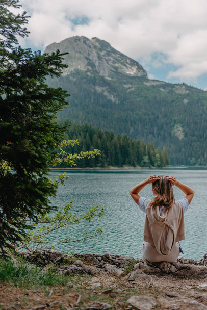Young woman on shore of mountain lake. The back of girl against background of mountains. Young girl sitting near by beautiful Black Lake in Montenegro. Original wallpaper from summer morning - Fotó, kép