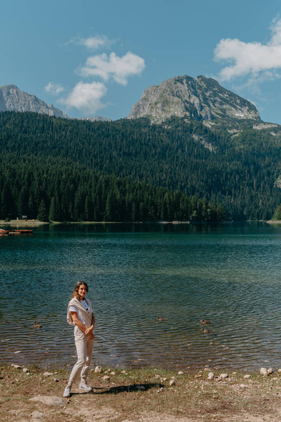 Girl looking to the lake, forest and mountains in Montenegro Durmitor national park. Tourist girl enjoys the magical view of the lake, coniferous forest and magical view sitting on big stone on the shore of a turquoise lake in the mountains. Hiking i - Foto, imagen