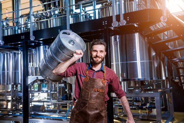 Young male brewer in leather apron holds barrel with craft beer at modern brewery factory - Foto, afbeelding