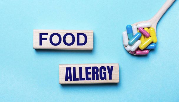On a light blue background, bright multi-colored pills in a spoon and two wooden blocks with the text FOOD ALLERGY. Medical concept - Fotó, kép