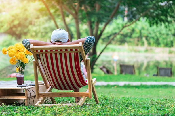 Rückenansicht eines älteren Mannes mit weißem Hut, der auf einem Gartenstuhl und am Tisch im Garten sitzt. Sommerurlaub im Grünen. Glückliche Person im Freien entspannen auf Liegestühlen im Garten.Freizeit im Freien - Foto, Bild