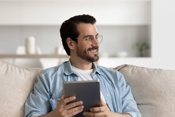 Happy millennial man in glasses resting on couch, using tablet - Фото, зображення