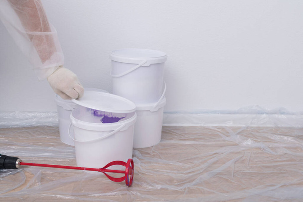 a construction worker's hand in protective clothing opens a bucket of paint to stir it, against a white wall - Valokuva, kuva