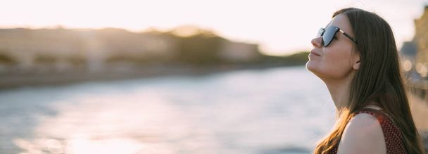 Портрет красивой молодой женщины на набережной реки на закате лета. Вечером девушка гуляет в европейском городке у реки на закате - Фото, изображение