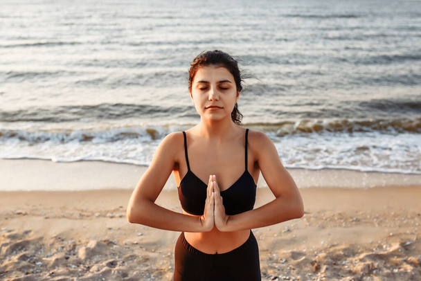 A girl with closed eyes in sportswear practices yoga. A close-up shot of a young woman against the backdrop of the sea or ocean - Foto, Imagen
