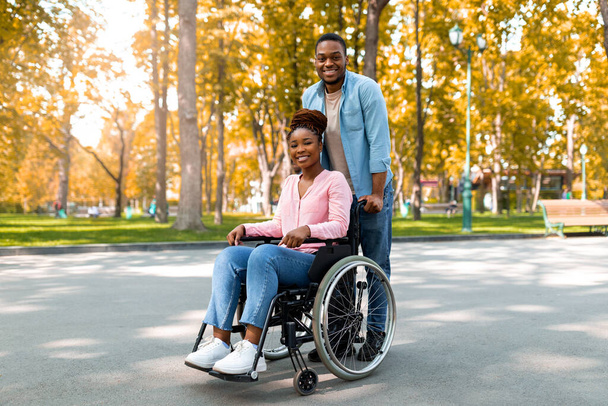Volle Länge des fröhlichen Schwarzen, der im Herbst seine behinderte Freundin im Rollstuhl im Stadtpark spazieren führt - Foto, Bild