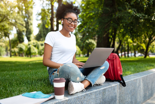 gelukkig zwart vrouw student met behulp van laptop dragen oordopjes zitten buiten - Foto, afbeelding