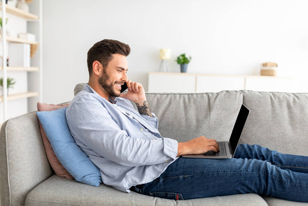 Koncept domácí kanceláře. Millennial businessman mluví na smartphone a pracuje na notebooku, sedí na gauči - Fotografie, Obrázek