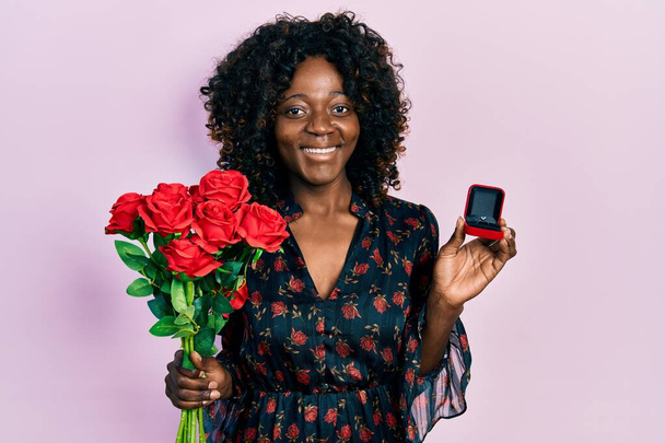Joven mujer afroamericana sosteniendo ramo de flores y anillo de compromiso guiño mirando a la cámara con expresión sexy, cara alegre y feliz.  - Foto, imagen