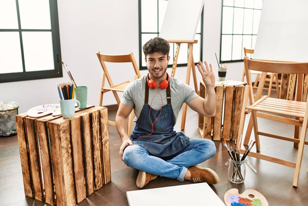 Молодий іспаномовний чоловік сидить у художній студії, показуючи і вказуючи пальцями номер чотири, посміхаючись впевнено і щасливо
.  - Фото, зображення