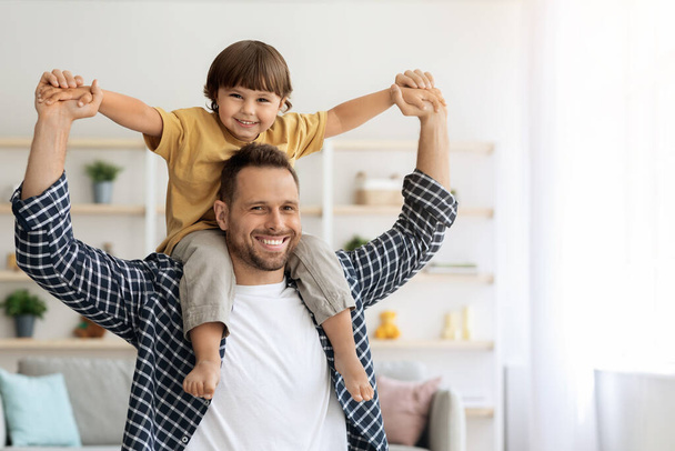 Cheerful father riding his little son on shoulders, having fun together at home, both smiling to camera, empty space - Photo, image