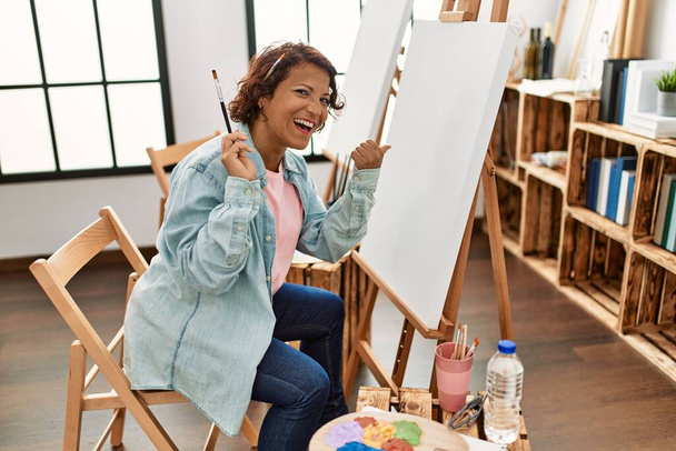 Middle age hispanic woman at art studio painting on canvas pointing thumb up to the side smiling happy with open mouth  - Фото, изображение