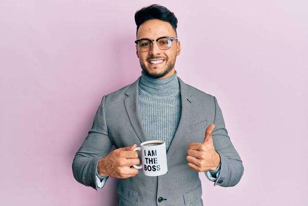 Joven árabe bebiendo de soy el jefe taza de café sonriendo feliz y positivo, pulgar hacia arriba haciendo excelente y signo de aprobación  - Foto, imagen