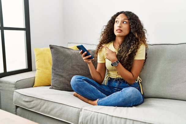 Hermosa mujer hispana sentada en el sofá de su casa usando un smartphone señalando a un lado preocupada y nerviosa con el dedo índice, preocupada y sorprendida expresión  - Foto, imagen