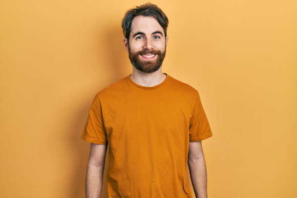 Hombre caucásico con barba vistiendo una camiseta amarilla casual con una sonrisa alegre y fresca en la cara. persona afortunada.  - Foto, Imagen