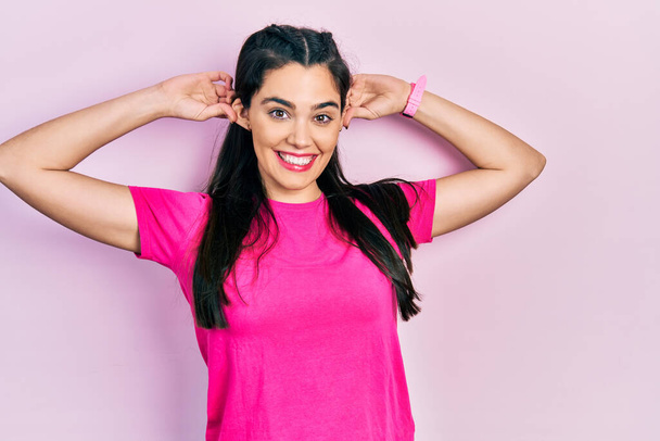 Молода іспаномовна дівчина в повсякденній рожевій сорочці посміхається тягнути вуха пальцями, смішний жест. проблема прослуховування
  - Фото, зображення