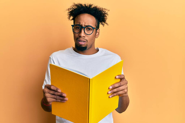 Ein junger afrikanisch-amerikanischer Mann mit Bart liest ein Buch und trägt eine Brille mit ahnungslosem und verwirrtem Gesichtsausdruck. Zweifelhaftes Konzept.  - Foto, Bild