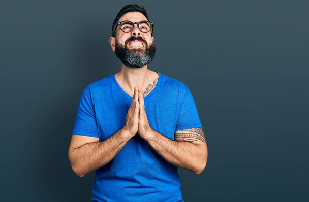 Hispanic man with beard wearing casual t shirt and glasses begging and praying with hands together with hope expression on face very emotional and worried. begging.  - Foto, imagen