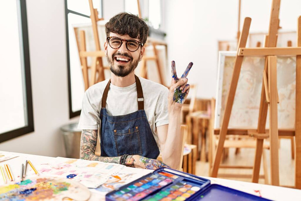 Ein spanischer Mann mit Bart im Kunststudio lächelt mit glücklichem Gesicht und zwinkert in die Kamera, als er ein Siegeszeichen macht. Nummer zwei.  - Foto, Bild