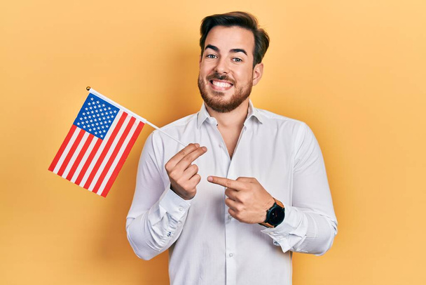 Knappe blanke man met baard die de vlag van de Verenigde Staten vasthoudt en vrolijk wijst met hand en vinger  - Foto, afbeelding