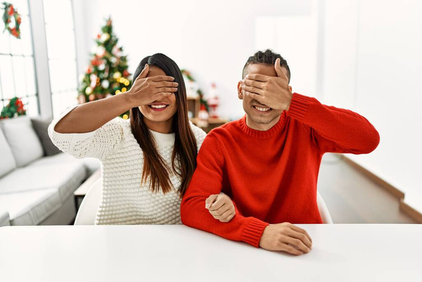 Masada oturan genç latin çift Noel ağacının yanında gülümseyerek ve gülerek gözleri kapayarak sürpriz yaptılar. kör kavram.  - Fotoğraf, Görsel