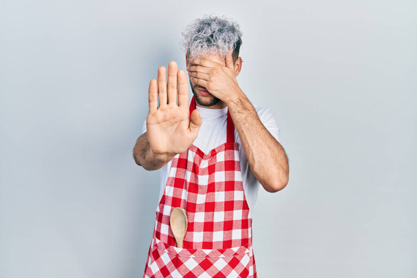 Young hispanic man with modern dyed hair wearing apron covering eyes with hands and doing stop gesture with sad and fear expression. embarrassed and negative concept.  - Photo, Image