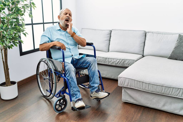 Bonito homem idoso sentado em cadeira de rodas na sala de estar tocando a boca com a mão com expressão dolorosa por causa de dor de dente ou doença dentária nos dentes. dentista  - Foto, Imagem