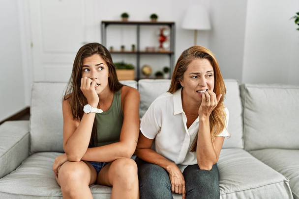 Äiti ja tytär istuvat yhdessä sohvalla kotona näyttäen stressaantuneilta ja hermostuneilta kädet suulla puremalla kynsiä. ahdistuneisuusongelma.  - Valokuva, kuva