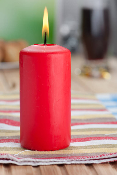 Płonąca czerwona świeca stoi na obrusie na drewnianym stole. - Zdjęcie, obraz