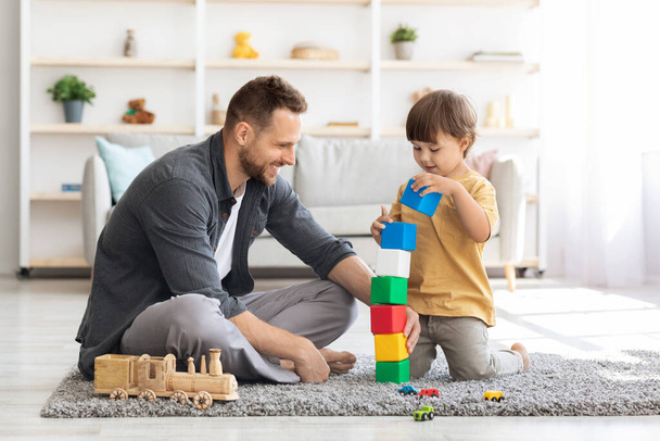 Spielzeit. Fröhlich liebender Vater spielt mit seinem kleinen Sohn bunte Blöcke, Junge baut zu Hause großen Turm - Foto, Bild