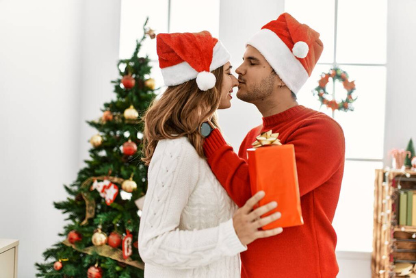 Young hispanic couple wearing christmas hat holding gift at home. - Fotó, kép
