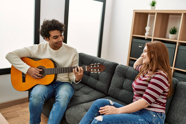 Junges Paar lächelt glücklich und spielt klassische Gitarre zu Hause auf dem Sofa. - Foto, Bild