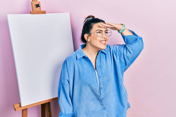 Young hispanic woman standing by painter easel stand very happy and smiling looking far away with hand over head. searching concept.  - Fotoğraf, Görsel