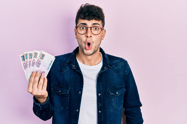 Young hispanic man holding egyptian pounds banknotes scared and amazed with open mouth for surprise, disbelief face  - Foto, Imagem