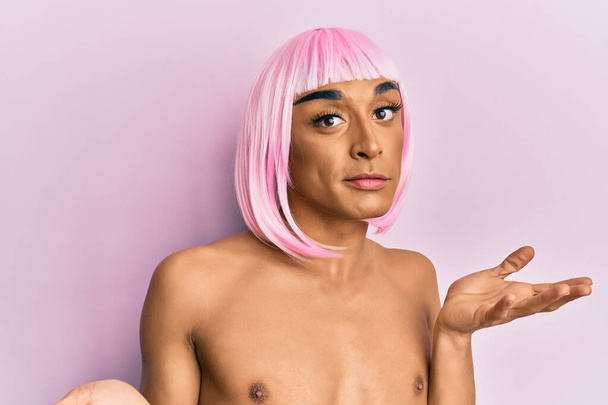 Hispanic transgender man wearing make up and pink wig clueless and confused expression with arms and hands raised. doubt concept.  - Foto, afbeelding