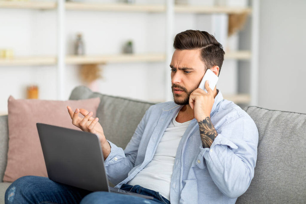 Hombre millennial descontento hablando por teléfono y mirando a la computadora portátil, teniendo problemas con la conexión a Internet, espacio de copia - Foto, Imagen