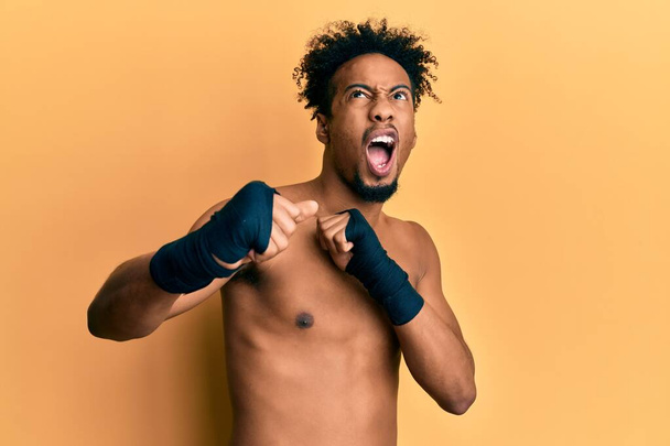 Young african american man with beard wearing boxing protection angry and mad screaming frustrated and furious, shouting with anger looking up.  - Photo, Image