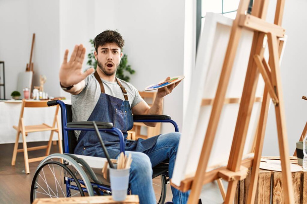 Jeune homme hispanique assis sur la peinture en fauteuil roulant au studio d'art faisant geste d'arrêt avec les paumes des mains, expression de colère et de frustration  - Photo, image