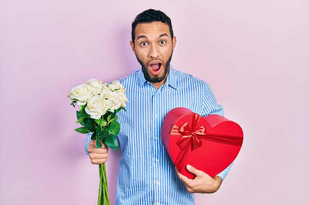 Homme hispanique avec barbe tenant cadeau d'anniversaire et bouquet de fleurs effrayé et choqué par l'expression surprise et étonnante, la peur et le visage excité.  - Photo, image