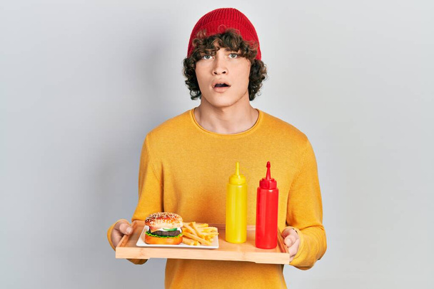 Красивый молодой человек ест вкусный классический бургер с кетчупом и горчицей в шоковом лице, выглядит скептически и саркастично, удивленный открытым ртом  - Фото, изображение