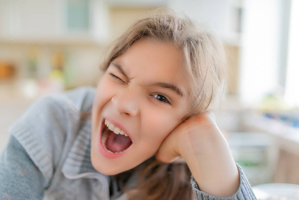 Uma menina adolescente loira bonita e alegre em uma camisola cinza boceja amplamente. Cozinha em segundo plano. - Foto, Imagem