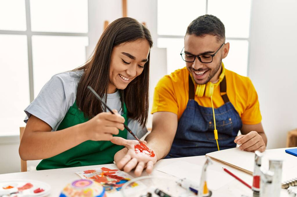 Молодая латиноамериканская пара улыбается счастливые руки живописи сидя на столе в художественной студии. - Фото, изображение