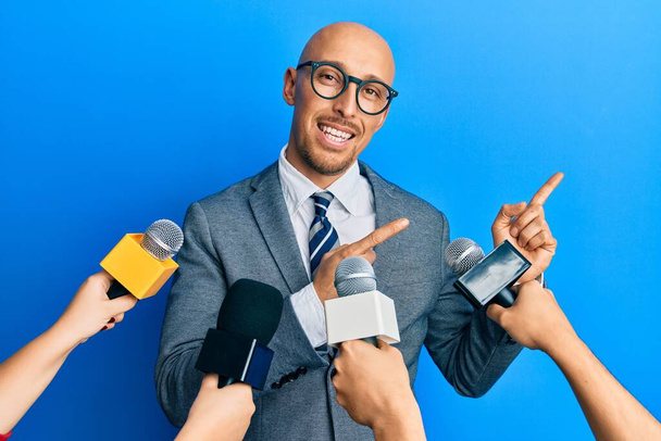 Homme chauve avec barbe interviewé par des journalistes tenant des microphones souriant et regardant la caméra pointant avec deux mains et doigts sur le côté.  - Photo, image