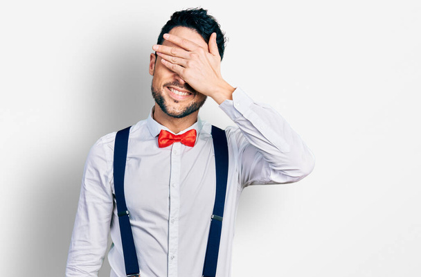 Hispanic man with beard wearing hipster look with bow tie and suspenders smiling and laughing with hand on face covering eyes for surprise. blind concept.  - Photo, Image