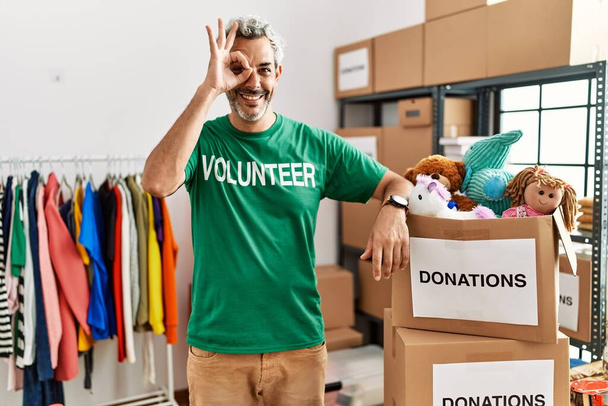Hombre hispano de mediana edad vistiendo camiseta voluntaria en el stand de donaciones haciendo buen gesto con la mano sonriente, los ojos mirando a través de los dedos con cara feliz.  - Foto, imagen