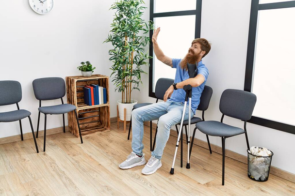 Jeune rousse homme utilisant des béquilles assis sur la chaise à la salle d'attente de la clinique. - Photo, image