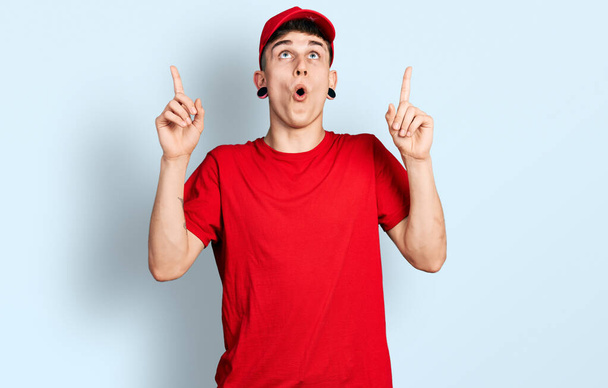 Jeune garçon caucasien avec dilatation des oreilles portant l'uniforme de livraison et le chapeau étonné et surpris regardant vers le haut et pointant avec les doigts et les bras levés.  - Photo, image